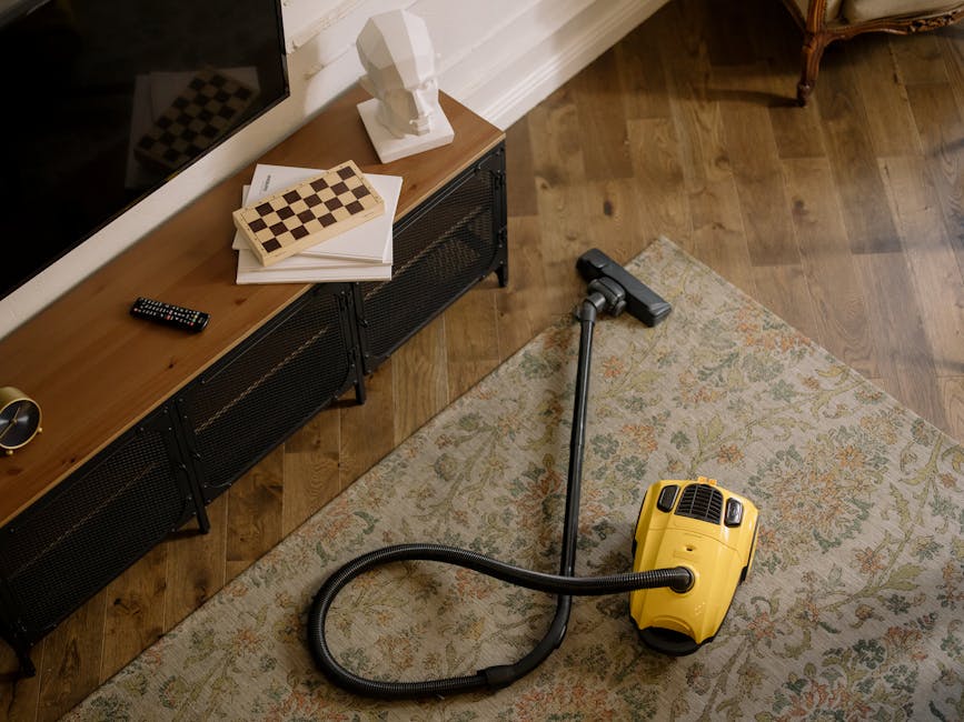 A yellow canister vacuum cleaner with a black hose and upright nozzle is positioned on a patterned area rug in a living room. The room features wooden flooring and a low, black metal and wood media co
