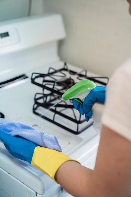 A person wearing blue and yellow cleaning gloves is holding a green and blue spray bottle and a cloth, standing in front of a white gas stove with black metal burners in a kitchen. The stovetop surface appears clean and free of dust or debris, with a beige or light-colored wall in the background. The lighting is natural or evenly distributed, highlighting the surface's cleanliness. This image illustrates the process of surface cleaning and sanitisation, integral to domestic cleaning services offered by Balham Cleaner, as described in the Balham High Road domestic cleaning guide for homeowners.