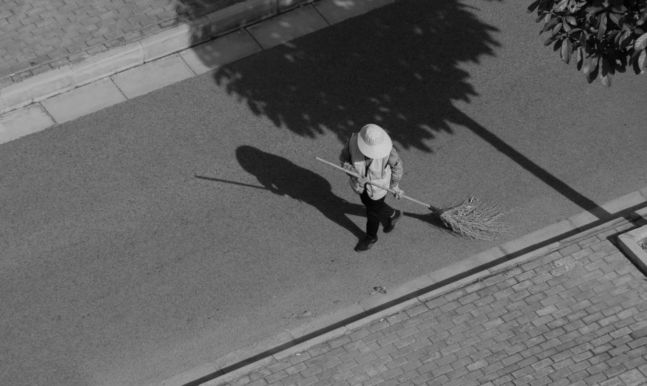 A person wearing a wide-brimmed hat and casual clothing is sweeping an outdoor paved pathway with a traditional straw broom, casting a distinct shadow on the ground. The pathway, part of a residential or commercial exterior, is bordered by a low brick wall and a section of patterned brick paving. Nearby, a tree provides shade, creating contrasting light and shadow on the surface. The scene emphasizes outdoor surface cleaning and maintenance, consistent with the focus of the Balham High Road domestic cleaning guide for homeowners by BALHAM Cleaner.