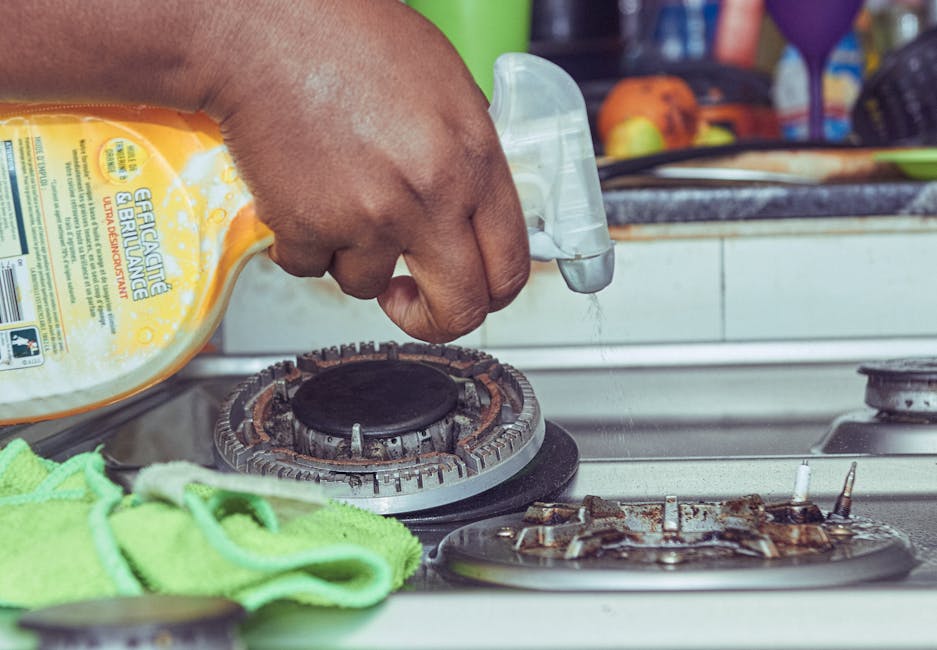 Close-up of a person wearing blue disposable gloves using a white cleaning cloth to wipe a smooth, light-colored surface, possibly a table or countertop, with a spray bottle of disinfectant cleaner he