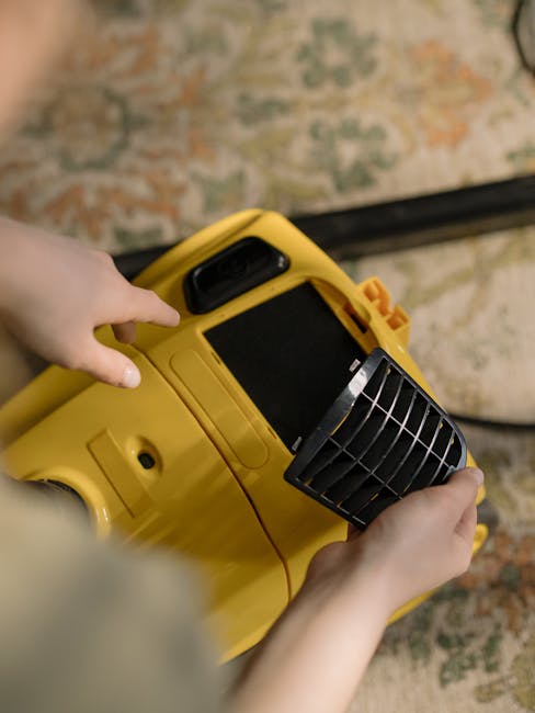 A yellow canister vacuum cleaner with a black hose and metal extension wand is positioned on a patterned area rug in a room with wooden flooring. Next to the vacuum, a light-colored wooden sideboard w